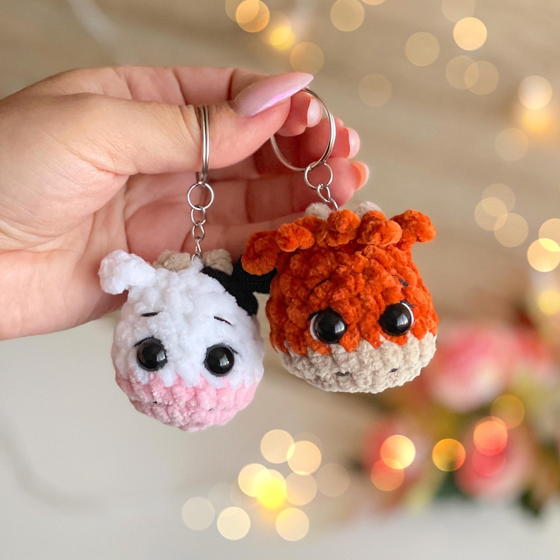 May include: Two handmade crochet keychains. One is a white cow with black spots and a pink muzzle, the other is an orange fox. Both have large black eyes and are attached to silver keyrings.