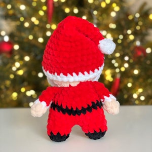 May include: A crocheted Santa Claus doll made of soft red, white, and black yarn. The figure wears a red hat with a white pom-pom, a red jacket with white trim, a black belt, and black boots. The doll has light beige yarn hands.
