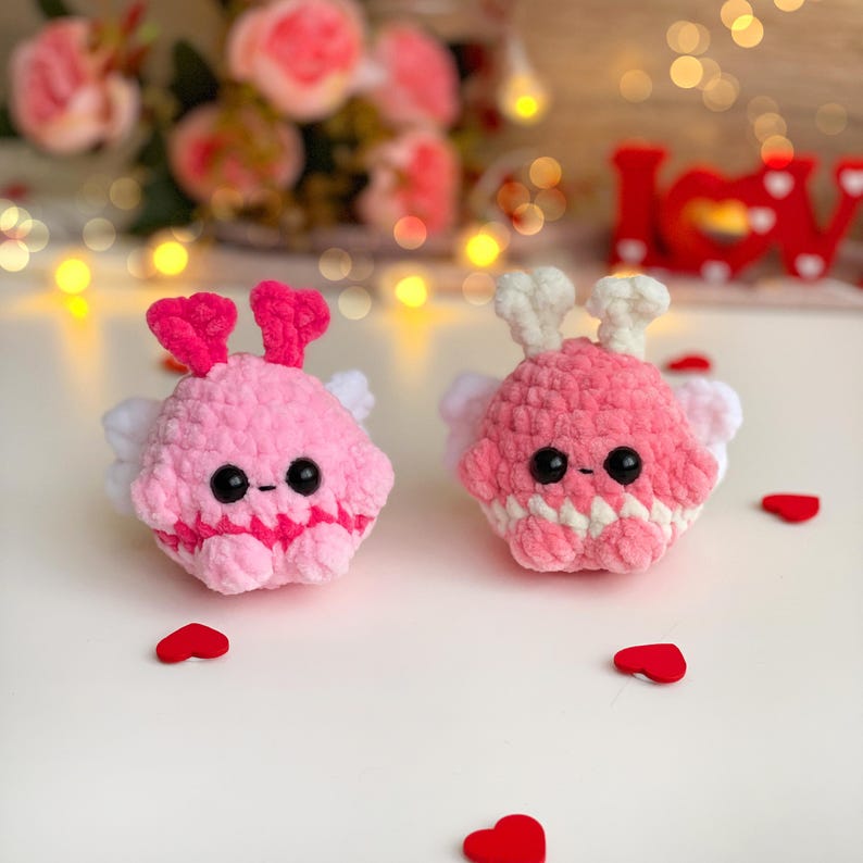 Peut inclure: Deux cr&eacute;atures rondes en crochet pelucheux avec des textures en laine rose, des yeux en perles noires et de petites ailes blanches. La figurine de gauche a des antennes rose vif, celle de droite des antennes blanches. Bouches brod&eacute;es.