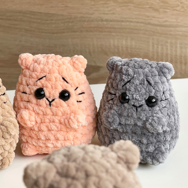 May include: Three crocheted plush toys shaped like cats with different colored fur. The cat on the left is light brown, the cat in the middle is peach, and the cat on the right is gray.