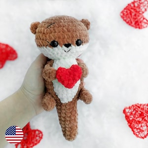 May include: A brown and white crocheted otter holding a red crocheted heart. The otter is sitting on a white surface with red hearts in the background.