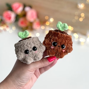 May include: Two small, plush, crocheted figures shaped like potted plants. One is light beige and the other is dark brown. Both have fuzzy textures, black plastic safety eyes, tiny embroidered mouths, pink blush marks, and a small green sprout on top.