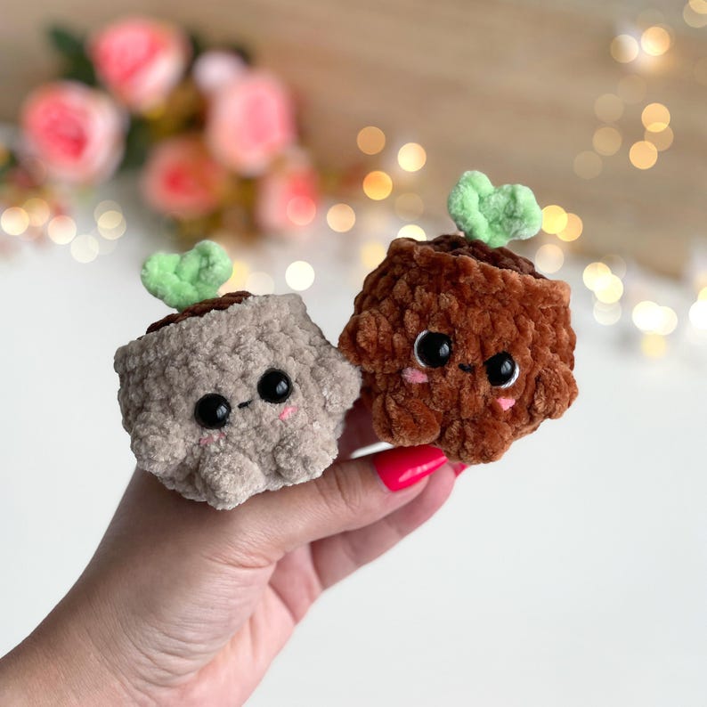 May include: Two small, plush, crocheted figures shaped like potted plants. One is light beige and the other is dark brown. Both have fuzzy textures, black plastic safety eyes, tiny embroidered mouths, pink blush marks, and a small green sprout on top.