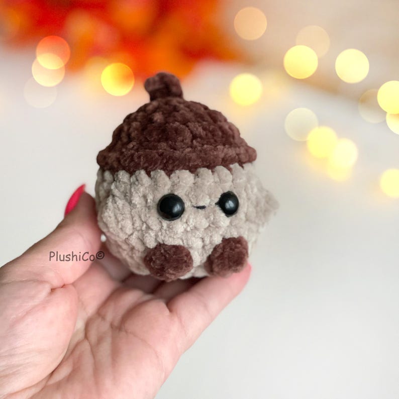 May include: A handmade plush acorn toy with a brown cap and a beige body. The acorn has two black button eyes and small brown feet. The toy is held in a hand, with the brand name "PlushiCo" visible.