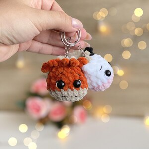 May include: Two handmade crochet keychains. One is orange and tan, resembling a fox, and the other is white with a pink nose and black accents. Both are attached to silver keyrings. The background is blurred with bokeh lights and flowers.