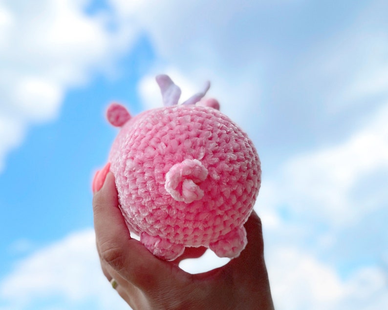 K&ouml;nnte beinhalten: Ein rosa geh&auml;keltes Schweinchenspielzeug mit einer wei&szlig;en Schleife auf dem Kopf. Das Schweinchen wird in einer Hand vor einem blauen Himmel mit wei&szlig;en Wolken gehalten.