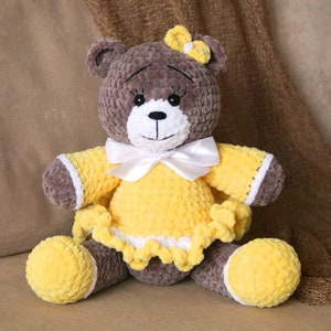 May include: A brown crocheted teddy bear wearing a yellow dress with white trim and a white bow. The bear has black eyes and a white nose.