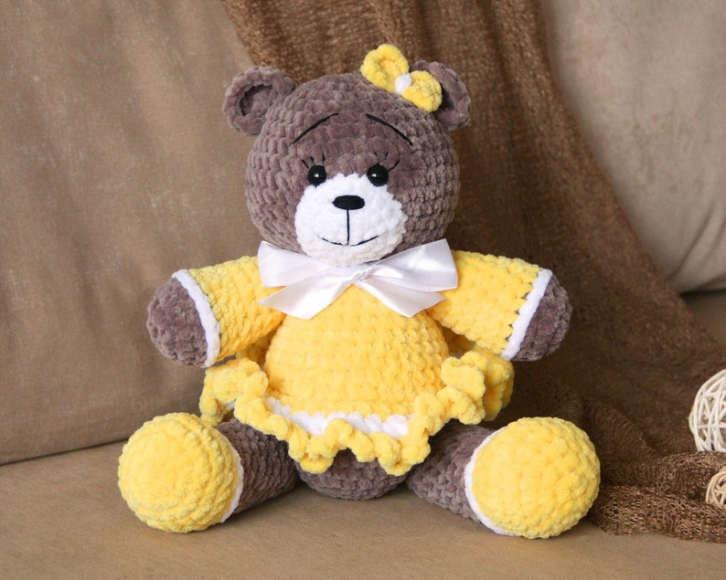 May include: A brown crocheted teddy bear wearing a yellow dress with white trim and a white bow. The bear has black eyes and a white nose.