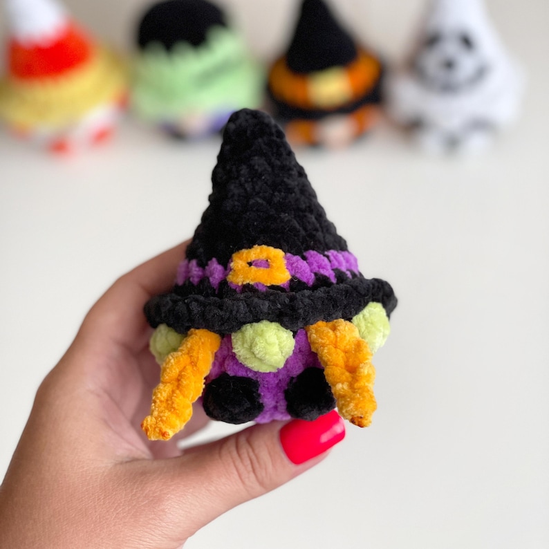 May include: A small, plush, crocheted witch figurine made of soft black, purple, orange, and lime green yarn. The figure features a tall, pointed black hat with a purple band and an orange buckle, orange yarn hair, and a round purple body.