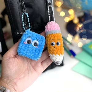 May include: Two handmade crochet keychains. One is a blue square with googly eyes and a sad face. The other is a pencil with orange, pink, gray, and white yarn, also with googly eyes. Both have small chain attachments.
