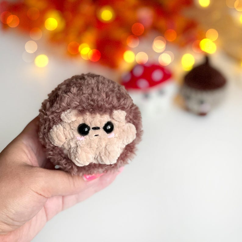 May include: A handmade, plush hedgehog with a light brown face and dark brown fur. The hedgehog has black eyes and a stitched mouth. In the background, there is a red mushroom with white spots and a brown acorn.