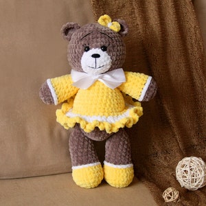 May include: A brown crocheted teddy bear wearing a yellow dress with white trim and a white bow. The bear is sitting on a brown fabric surface.