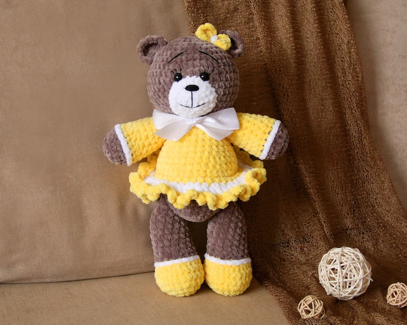 May include: A brown crocheted teddy bear wearing a yellow dress with white trim and a white bow. The bear is sitting on a brown fabric surface.