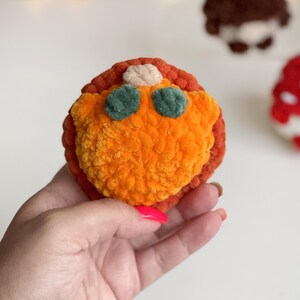 May include: A round, plush, crocheted object featuring a textured orange center, a dark orange outer border, two small dark green circular accents, and a small beige top detail. The item is crafted from soft, fuzzy yarn with a visible stitch pattern.