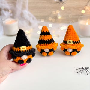 May include: Three small crocheted gnomes featuring pointed hats in black and orange yarn. One gnome wears a solid black hat with an orange band, while the other two feature orange and black striped patterns. Each gnome has a small beige nose.