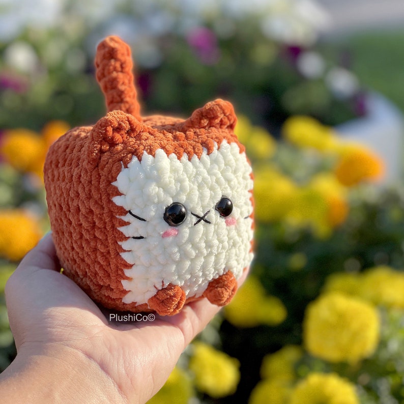 May include: A crocheted plush toy shaped like a cat with orange and white fur. The cat has black eyes and a cute expression.