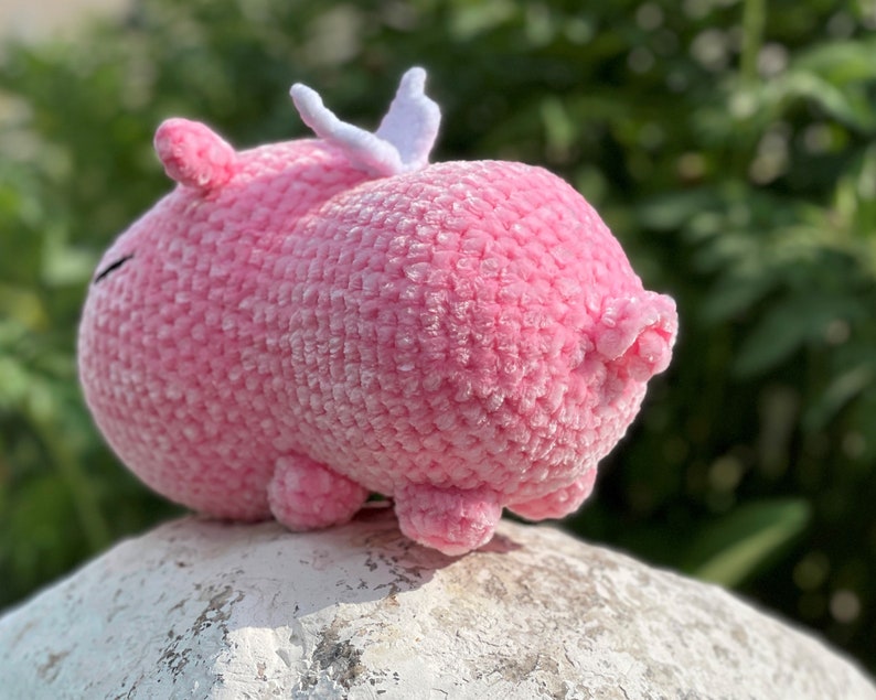 K&ouml;nnte beinhalten: Ein rosa geh&auml;keltes Schwein mit wei&szlig;en Fl&uuml;geln. Das Schwein sitzt auf einem wei&szlig;en Stein.