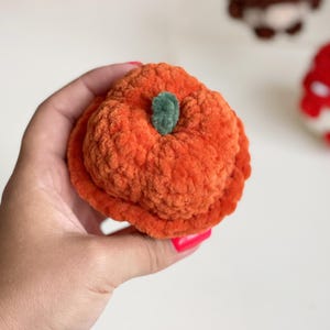 May include: A small, round, orange crocheted pumpkin made from soft, plush yarn. The pumpkin features a textured, ribbed surface and a small, dark green crocheted stem centered on top. The object is held in a hand, showcasing its compact size.