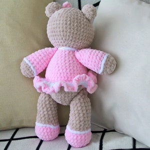 May include: A crocheted teddy bear wearing a pink dress with white trim and a white ruffle. The bear is standing on a checkered surface.