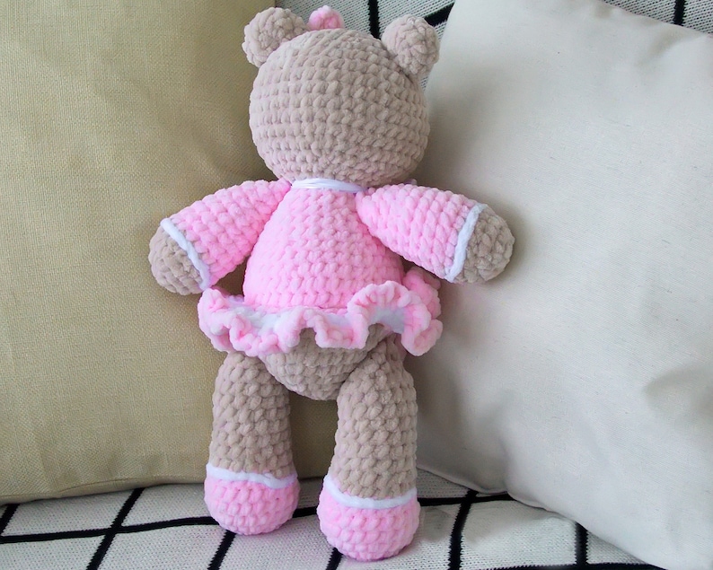May include: A crocheted teddy bear wearing a pink dress with white trim and a white ruffle. The bear is standing on a checkered surface.
