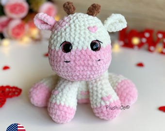 May include: A crocheted pink and white cow plush toy with a heart on its forehead and sparkly eyes. The toy is sitting on a white surface with red hearts scattered around it.