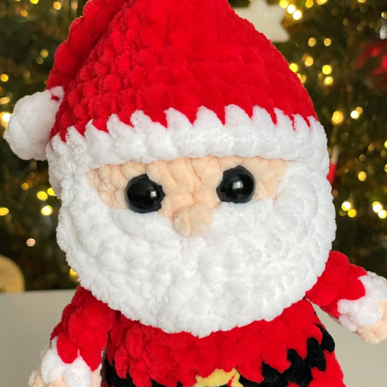 May include: A plush Santa Claus doll crafted from soft red and white yarn. The figure features a red hat with a white pom-pom, a thick white beard, a peach-colored face, and large black safety eyes. A black belt with a yellow buckle is visible.