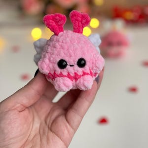 Peut inclure: Une petite peluche crochet&eacute;e rose, ronde et textur&eacute;e. Elle pr&eacute;sente deux antennes roses en forme de c&oelig;ur, deux petites ailes blanches sur les c&ocirc;t&eacute;s, ainsi que deux yeux en plastique noir et une petite bouche brod&eacute;e.