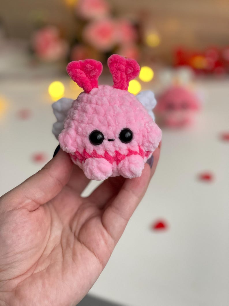 Peut inclure: Une petite peluche crochet&eacute;e rose, ronde et textur&eacute;e. Elle pr&eacute;sente deux antennes roses en forme de c&oelig;ur, deux petites ailes blanches sur les c&ocirc;t&eacute;s, ainsi que deux yeux en plastique noir et une petite bouche brod&eacute;e.