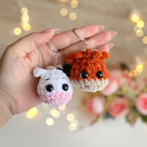 May include: Two handmade crochet keychains: a white cow with pink accents and a fluffy orange fox. Both have large black eyes and silver key rings. The cow keychain has black spots and the fox has a beige muzzle.