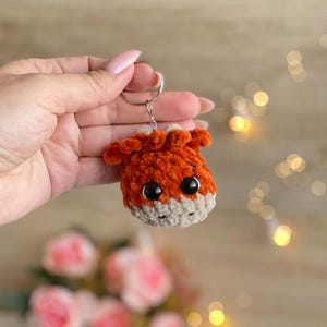 May include: A handmade orange and tan crochet keychain in the shape of a small animal head with large black eyes. The keychain is attached to a silver metal ring. The plush toy is held in a person's hand.