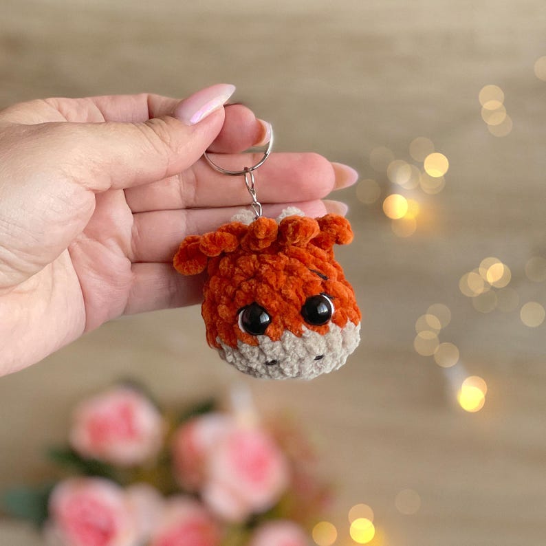 May include: A handmade orange and tan crochet keychain in the shape of a small animal head with large black eyes. The keychain is attached to a silver metal ring. The plush toy is held in a person's hand.