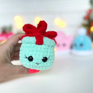 May include: A small, mint green crocheted plush toy shaped like a gift box. It features a bright red crocheted bow on top, two black plastic safety eyes with stitched eyelashes, and a small, simple stitched mouth on the front surface.