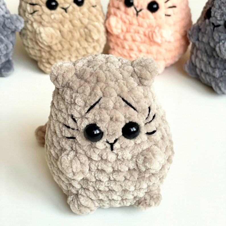 May include: A plush crocheted cat toy with a gray and brown color scheme. The cat has black eyes and a stitched-on black nose and mouth.