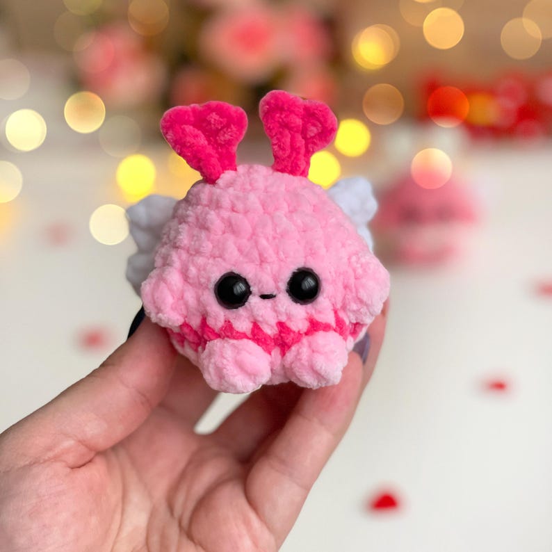 Peut inclure: Un petit jouet en peluche crochet&eacute;, rond et rose, avec une surface textur&eacute;e et duveteuse. Il pr&eacute;sente deux antennes roses dress&eacute;es, deux ailes blanches sur les c&ocirc;t&eacute;s, et deux grands yeux de s&eacute;curit&eacute; en plastique noir brillant avec une petite bouche brod&eacute;e.