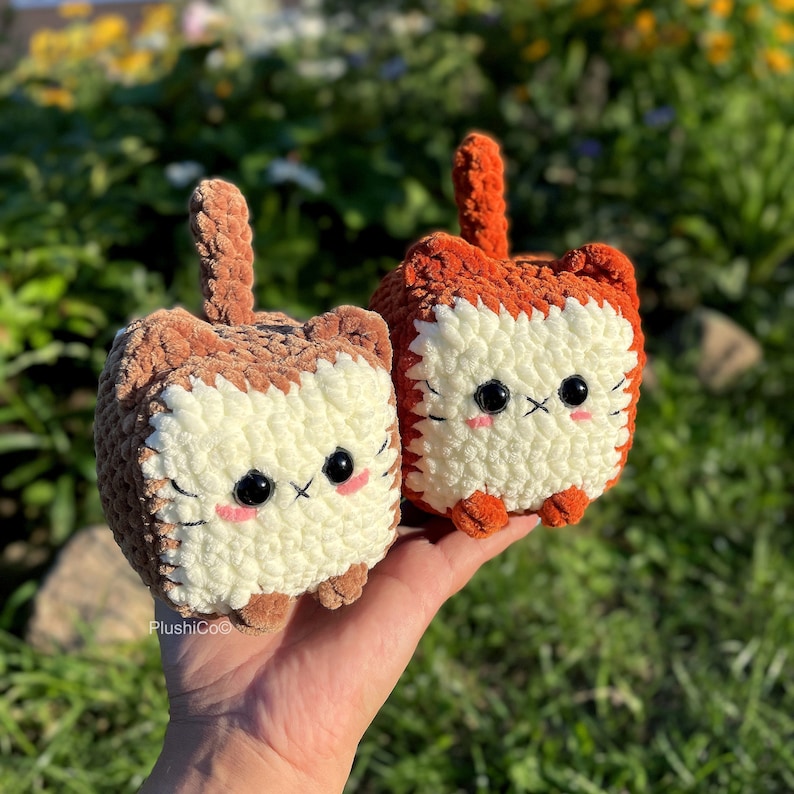 May include: Two crocheted plush cat toys, one brown and one orange, with white fur and black eyes. The toys are sitting on a hand.