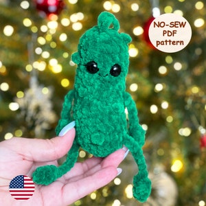 May include: A green crocheted pickle ornament with black eyes and a friendly face. The ornament is being held in a hand against a blurry background of a Christmas tree with lights. The text "NO-SEW PDF pattern" is visible in the upper right corner of the image.
