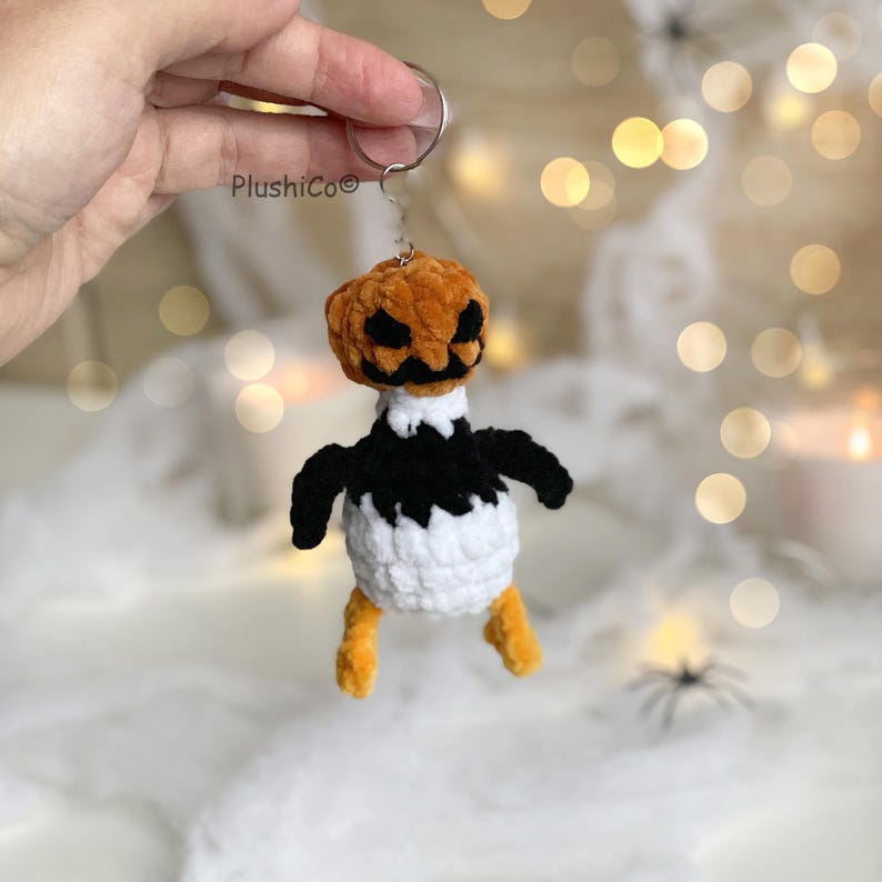 May include: A handmade Halloween-themed keychain featuring a crocheted pumpkin head with a carved face, atop a black and white body. The keychain has orange legs and black wings. The brand name "Plushico" is visible on the keychain.