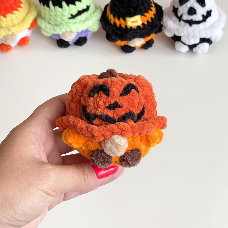 May include: A small, round, orange crochet pumpkin figure with a textured yarn surface. The pumpkin features a dark brown stem, a stitched black smiling face, and small brown feet. In the background, four other colorful crochet figures are visible.