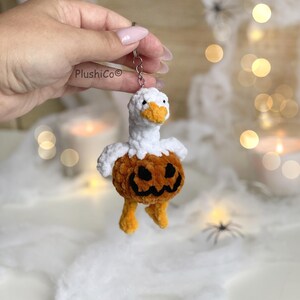 May include: A handmade keychain featuring a white goose with an orange beak and feet, and a pumpkin body with a carved jack-o'-lantern face. The plushie is attached to a silver keyring. The keychain is a Halloween-themed accessory.