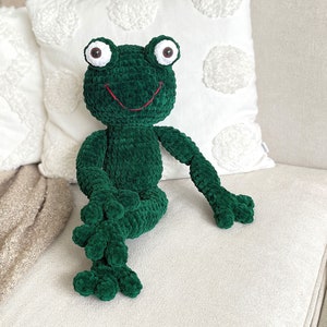 May include: A green crocheted frog plush toy with large brown eyes and a red embroidered smile. The frog is sitting on a white sofa.