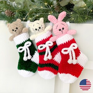 May include: Three crocheted Christmas stockings with different animal characters. A brown bear, a white cat, and a pink bunny are peeking out of the stockings. The stockings are decorated with white, red, and green stripes and white bows.