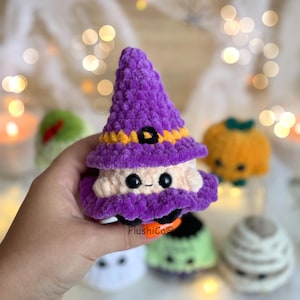 Puede incluir: Una mu&ntilde;eca de bruja de ganchillo con un sombrero morado, ribete naranja y hebilla negra. La bruja tiene una cara color crema con ojos negros y una peque&ntilde;a sonrisa. Otras mu&ntilde;ecas con tem&aacute;tica de Halloween son visibles en el fondo.