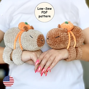 May include: Two crocheted stuffed capybaras wearing orange pumpkin hats. The capybaras are brown and beige and are wearing the hats as bracelets on a person's wrists. The text "Low-Sew PDF pattern" is visible above the capybaras.