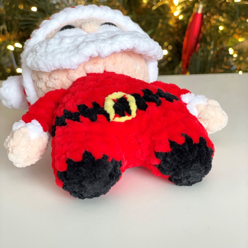 May include: A plush Santa Claus doll made of soft red, white, and black yarn. The figure features a white beard and hat trim, a red suit with a black belt, a yellow buckle, and black boots. The doll has light peach-colored hands and face.