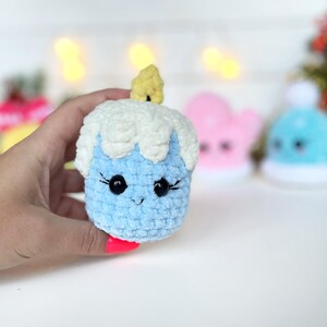 May include: A small, light blue crocheted plush toy shaped like a candle with white textured yarn at the top representing melting wax and a small yellow flame on top. The toy features two black plastic eyes with stitched eyelashes.