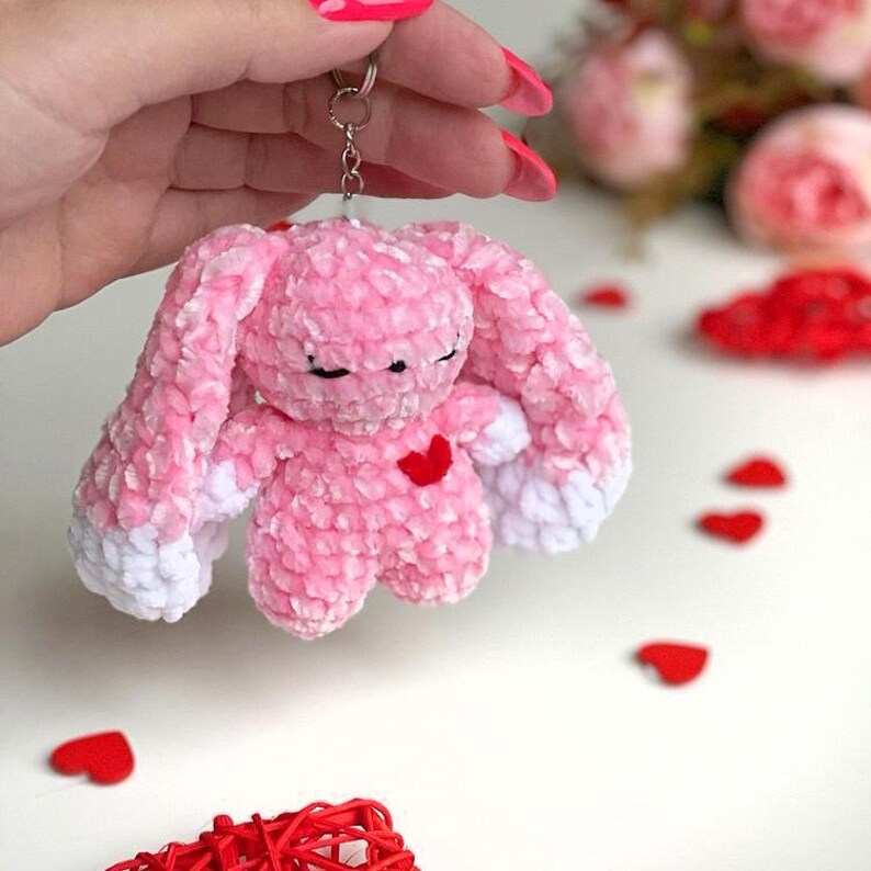 May include: Pink crocheted bunny keychain with white accents and a red heart on its chest. The bunny is holding its paws together and has closed eyes.