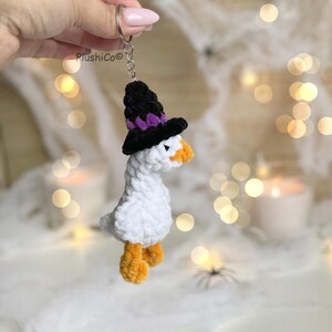 May include: A white plush goose keychain wearing a black witch hat with purple accents and orange feet and beak. The keychain is attached to a silver ring. The brand name "PlushiCo" is visible on the left.