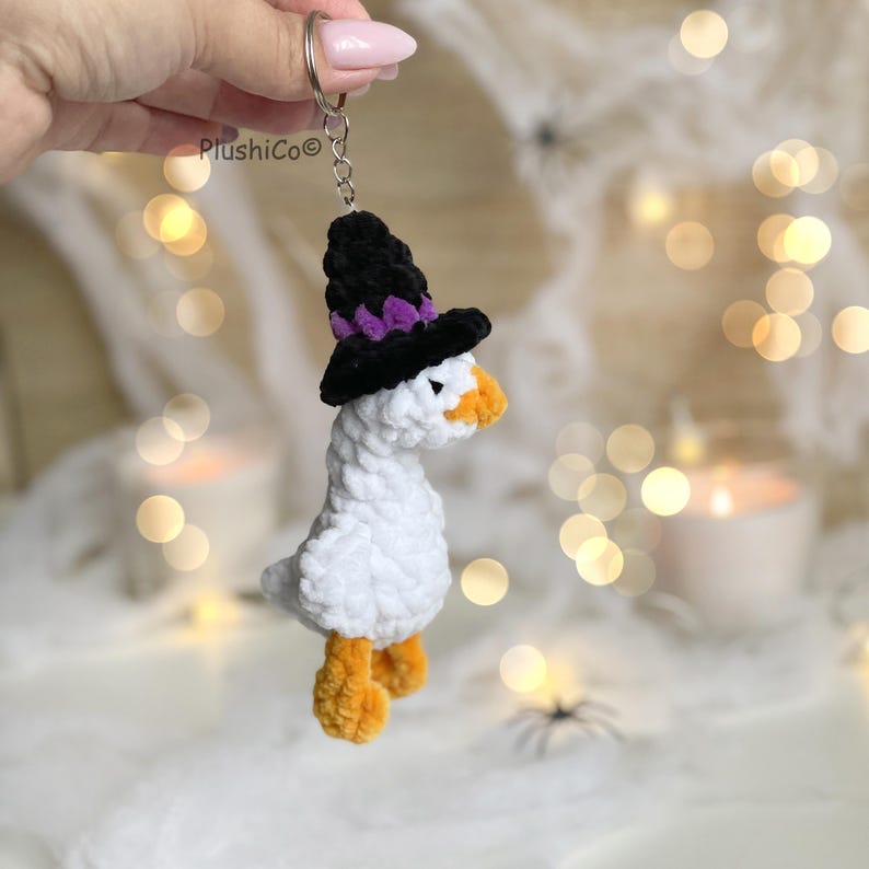 May include: A white plush goose keychain wearing a black witch hat with purple accents and orange feet and beak. The keychain is attached to a silver ring. The brand name "PlushiCo" is visible on the left.
