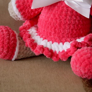 May include: A pink crocheted stuffed animal with a white bow and a white crocheted trim around the skirt. The animal is lying on a brown surface.