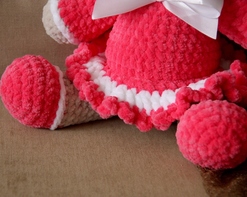 May include: A pink crocheted stuffed animal with a white bow and a white crocheted trim around the skirt. The animal is lying on a brown surface.